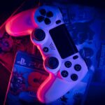 Image of a white PlayStation 4 controller resting on a stack of video game cases. The controller is illuminated in pink and blue, creating a striking contrast against the dark background. The cases feature a variety of titles, including "LEGO Jurassic World" and "PlayStation 5," suggesting a diverse collection of gaming options. The overall composition evokes a sense of excitement and anticipation for gaming adventures.