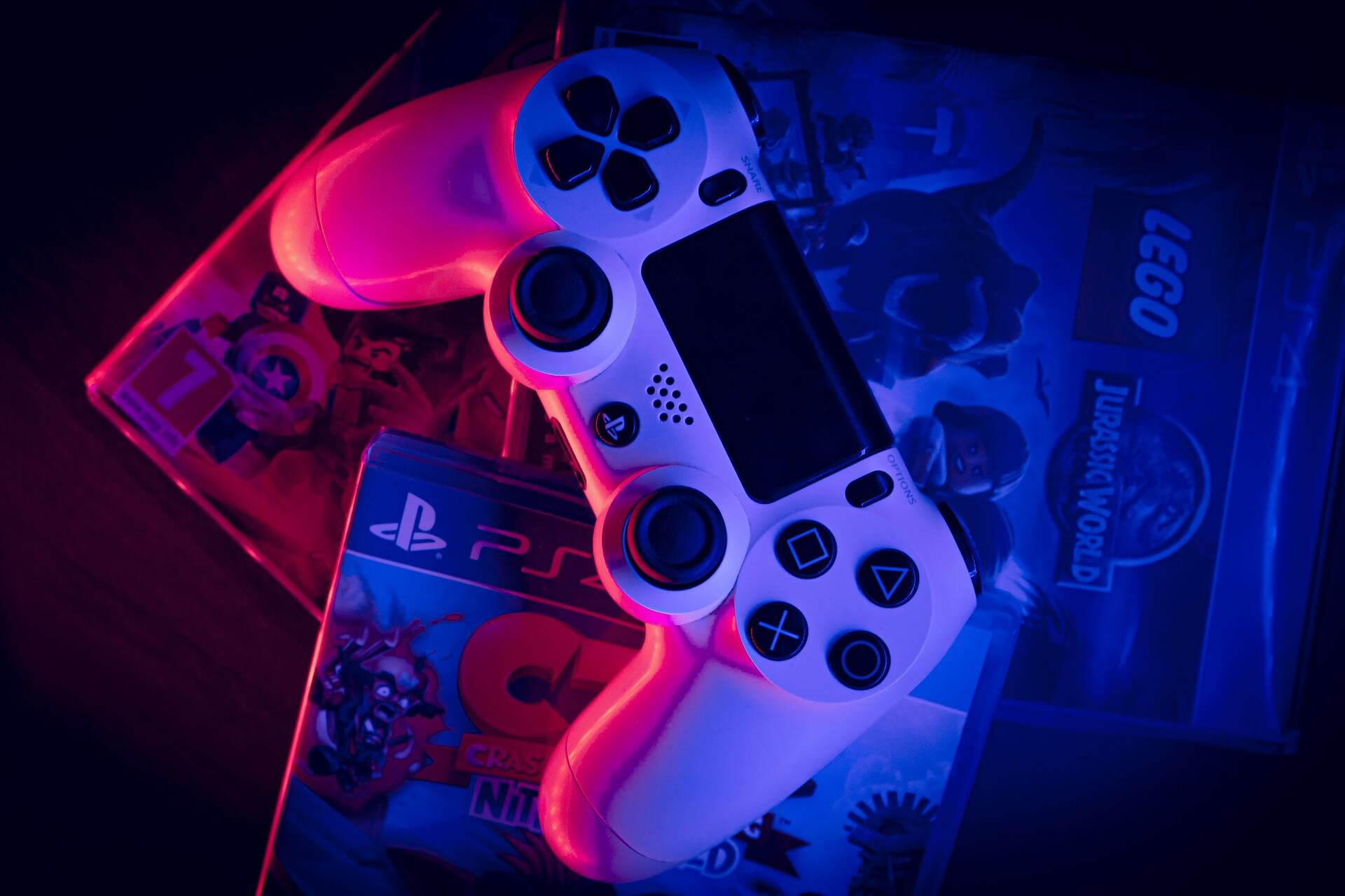 Image of a white PlayStation 4 controller resting on a stack of video game cases. The controller is illuminated in pink and blue, creating a striking contrast against the dark background. The cases feature a variety of titles, including "LEGO Jurassic World" and "PlayStation 5," suggesting a diverse collection of gaming options. The overall composition evokes a sense of excitement and anticipation for gaming adventures.